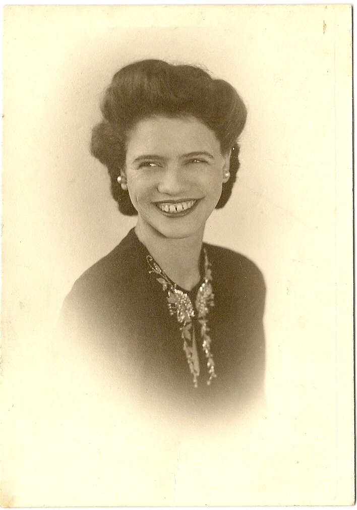 Aunt Mabel in a sepia studio portrait from the 1940s — dark waved hair, a wide open smile, and a sparkling floral brooch at the collar of her dark jacket.
