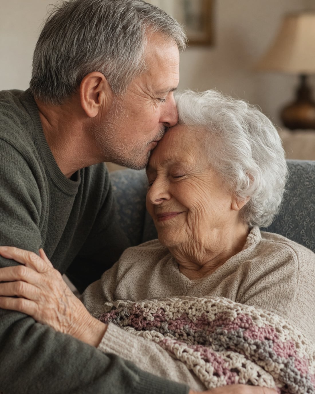 A son kisses his mother's forehead — she smiles, eyes closed, wrapped in a crocheted blanket.